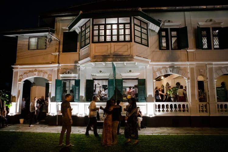 Na Café after dark. Landscape lighting subtly lights two people in the grass, hugging amidst people with drinks in hand walking, talking, standing and sitting on a bench. Many more people are visible on the entrance steps and inside the café through a trio of windows with security bars and green shutters and three archways.