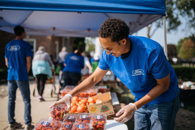 Male individual wearing the blue Ford Volunteer Corps T-shirt, places strawberries on a table with more strawberries and onions on additional tables in the background. Additional people in the background some we wearing Volunteer Corps T-shirts, others shopping.