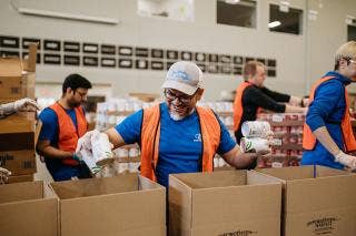 Male holding cans in both hands, placing items into Forgotten Harvest logoed boxes, surrounded by volunteers working in the warehouse