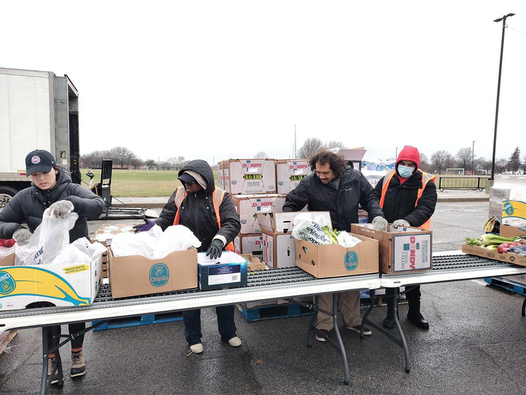 Four people, outdoors, bundled up wearing hooded jackets and hats and gloves filling produce boxes on a conveyor belt with celery and other food items.