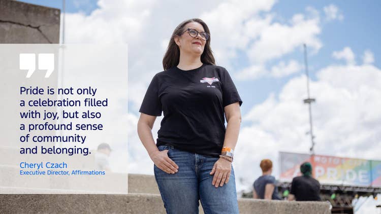 Female stands outdoors with a rainbow coloured PRIDE billboard in the background. She wears an Affirmation logoed T-shirt with the quote &quot;Pride is not only a celebration filled with joy, but also a profound sense of community and belonging.&quot; Cheryl Szach Executive Director, Affirmations
