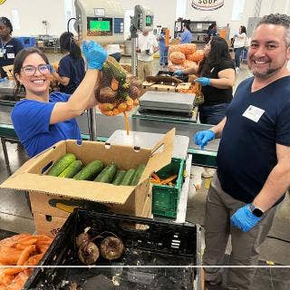 A female wearing a ponytail and glasses proudly holding a mesh bag filled with carrots, turnips and zucchini. In front of her is a big box of zucchini, some turnips, carrots. Across from her is a male with a small beard, looking at the camera. In the background are others weighing and packing boxes with vegetables.