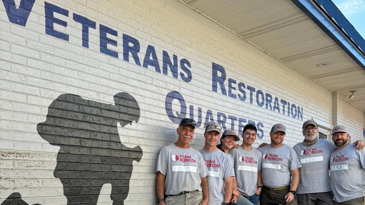 Seven people clad in Team Rubicon T-shirts stand in front of a building with a silhouette of a soldier taking a knee with head bowed and hands clasped together and VETERANS RESTORATION QUARTERS. At their feet is a U.S. flag on a sign with UNITED WE STAND GOD BLESS AMERICA.