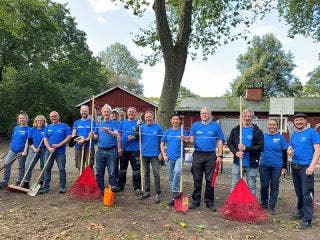 Thirteen people in blue Ford Philanthropy T-shirts, many holding rakes, brooms, shovels, levelers as they stand in line, outside, facing the camera with a crimson building in the background.