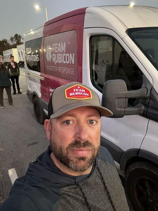 Bearded and mustached male wearing Tean Rubicon baseball cap beside TEAM RUBICON DISASTER RESPONSE vehicle.