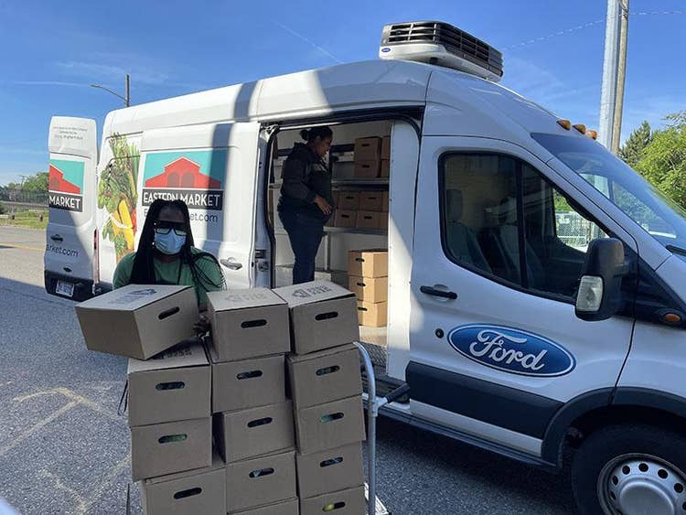 A hand truck filled with 15 cardboard boxes with the Eastern Market logo on the lids arranged by a masked person. A second person stands in the van surrounded by about the same number of bozes, inside an Eastern Market and Ford logoed Transit van.
