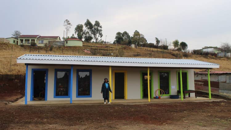 One story building with corrugated metal roof. The left doorway, two windows and two pillars are bright blue, a person stands camera left of the first of two yellow pillars with a centered yellow doorframe, a third doorway then two windows and two pillars are light lime green on the right end of the building. Behind the school building are sparsely scattered buildings with fenced areas and a few trees.