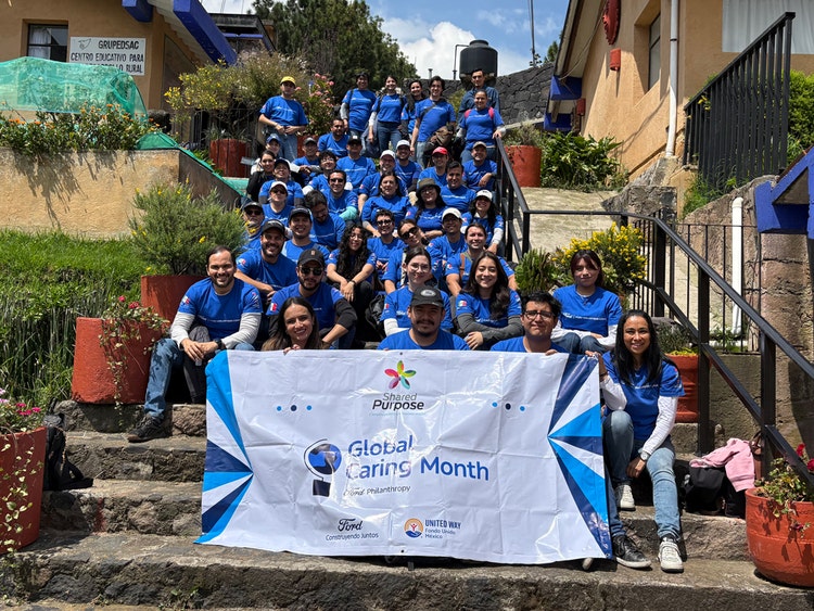 A group of people in matching Ford blue shirts sit and stand on outdoor steps, holding a banner that reads "Global Caring Month" with several logos and text.
