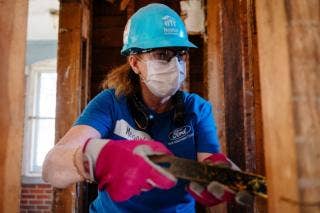 Woman holding a crowbar while next to house framing with gloves on and a hard hat
